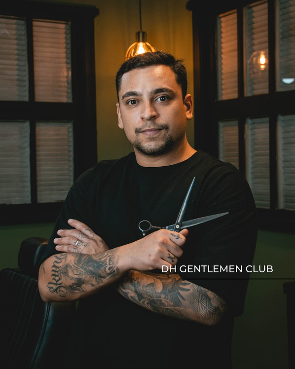 Foto do barbeiro Douglas de uniforme preto, posicionado no centro da barbearia com decoração rústica e com detalhes de uma janela com moldura em madeira ao fundo, realizando um corte de cabelo com tesoura em um cliente sentado na cadeira profissional.